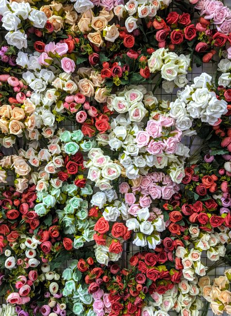 A dense display of artificial roses in various colors, including white, pink, red, peach, and light green, arranged in an overlapping pattern. The flowers have soft, velvety petals with detailed textu