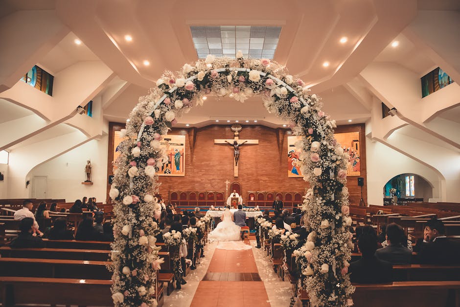 A large floral arch composed of white and blush pink flowers, including roses and hydrangeas, decorates the entrance aisle of a church. The arch is densely packed with blooms and greenery, creating a 