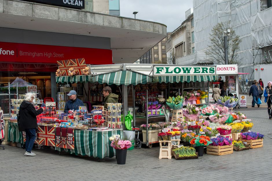 A street-side flower stall with a green and white striped canopy displaying a variety of fresh floral arrangements and bouquets in vibrant colors, including pink, purple, orange, and yellow, wrapped i