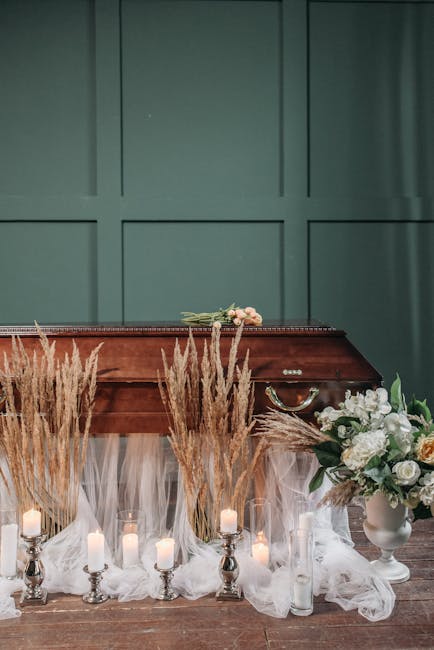 A floral tribute featuring a bouquet of soft pink roses and white lilies resting on a polished wooden casket, which is surrounded by tall, golden pampas grass arrangements and small white candles in s