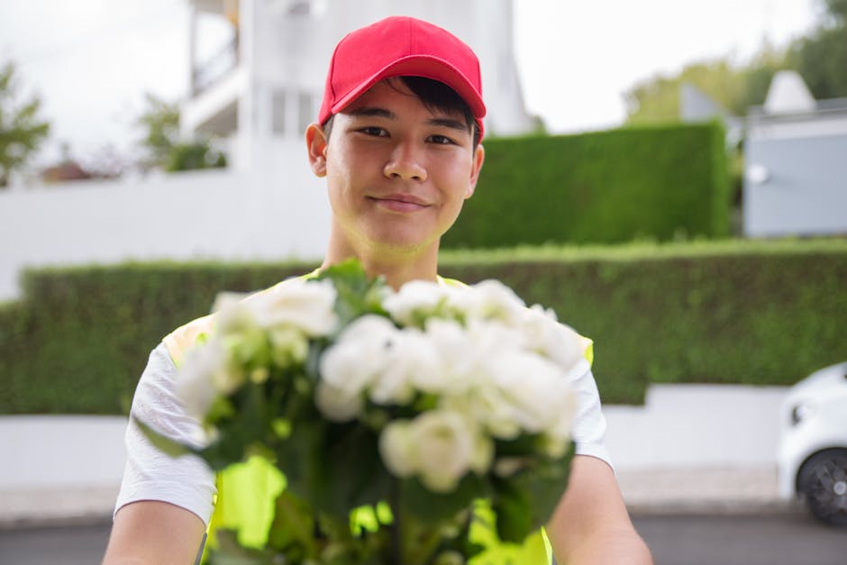 A delivery person in a red cap and high-visibility vest holds a bouquet of fresh white flowers, including roses and peonies, wrapped in clear plastic. The flowers are vibrant and appear recently picke