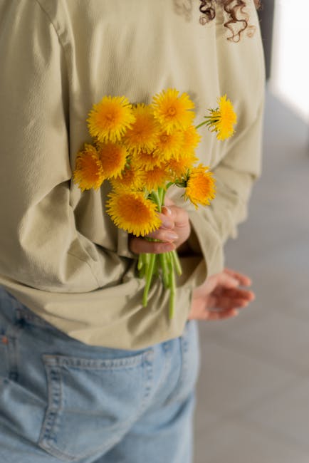 A person wearing a beige long-sleeve top and light blue jeans is holding a bouquet of bright yellow pom-pom-shaped flowers with green stems, assembled in a casual hand grip. The flowers appear fresh,